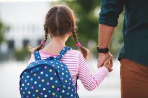 Photo d'une élève avec un sac à dos, tenant la main d'un adulte. Ils sont tous les deux de dos. 